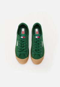 Green canvas sneakers with laces, featuring a gum rubber sole and a logo tag on the side. The inner sole displays branding with red and blue accents.