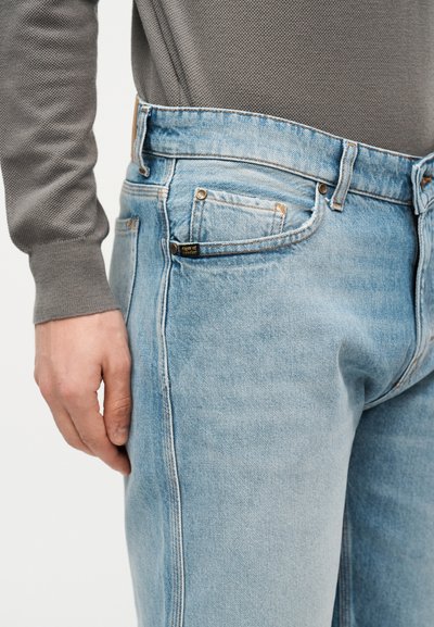 Άτομο που φοράει τζιν παντελόνι σε ανοιχτό μπλε και γκρι πουκάμισο με μακριά μανίκια, με το χέρι χαλαρό δίπλα στο σώμα, εμφανείς οι λεπτομέρειες από τις μπροστινές και πλαϊνές τσέπες.