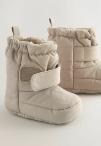 Bottines beiges rembourrées avec une doublure douce, une sangle velcro, un haut froncé et une semelle blanche texturée avec un motif subtil.