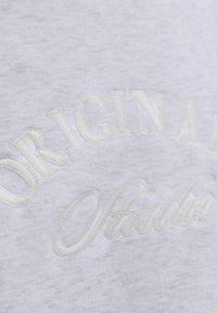 Lichtgrijze sweatshirt met geborduurde tekst "ORIGINAL" en "Studio" in een licht verhoogd, off-white draad. Zachte textuur en casual ontwerp.