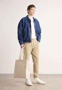 Jeffrey petroleumsjakk med glidelåslommer, hvit t-skjorte, beige bukser med elastisk mansjett og hvite joggesko. Beige totebag med to stropper.