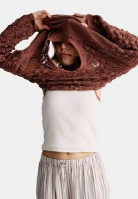 Pull en tricot marron à la coupe ouverte, avec un large col, porté par-dessus un débardeur blanc, en tissu doux et texturé.
