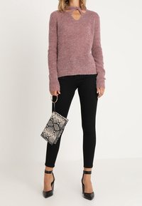 Vrouw in een mauve gebreid trui met een keyhole halslijn, zwarte skinny broek, zwarte enkelband-hakken, met een tas in slangenprint.