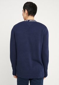 Tummansininen neulepaita, jossa on pitkät hihat, ribbireunus kauluksessa ja hihoissa. Paita sisältää kontrastivärisen lapun kaula-aukossa ja suoran helman.