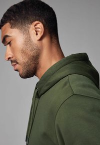 Sweat à capuche vert olive avec cordon de serrage, tissu texturé et coupe décontractée. Le profil latéral met en avant la forme de la capuche et les détails de couture.