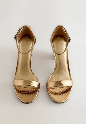 Kullatud kõrge kontsaga sandaalid, millel on tekstuuriga mudeleid sarnane nahk. Omab pahkluu rihma, kahte laia esiriba ja läikivat kuldset sisemust.