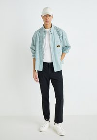 Chemise à rayures verte claire et blanche, portée sur un t-shirt blanc, associée à un pantalon noir et des baskets blanches. Le modèle porte une casquette beige.