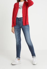 Rode cardigan over een wit t-shirt met print, gecombineerd met blauwe skinny jeans en witte sneakers. Soepele texturen en een casual ontwerp.