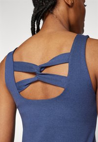 Widok z tyłu osoby noszącej niebieską bezrękawnik z skręconym detalem na górnej części pleców i plecionymi włosami.