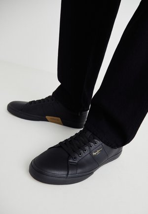 Black leather sneakers with a sleek design, featuring a textured surface, round toe, and black laces. Gold logo detail on the side.