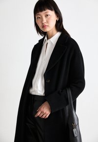 Langer schwarzer Wollmantel mit Reverskragen, doppelten Knöpfen und oversized Passform. Kombiniert mit einem weißen Hemd und schwarzen Hosen. Schwarze Handtasche.