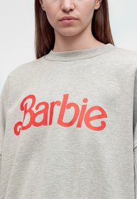 Jeune personne portant un sweat-shirt gris clair avec le mot "Barbie" imprimé en grandes lettres rouges sur la poitrine.