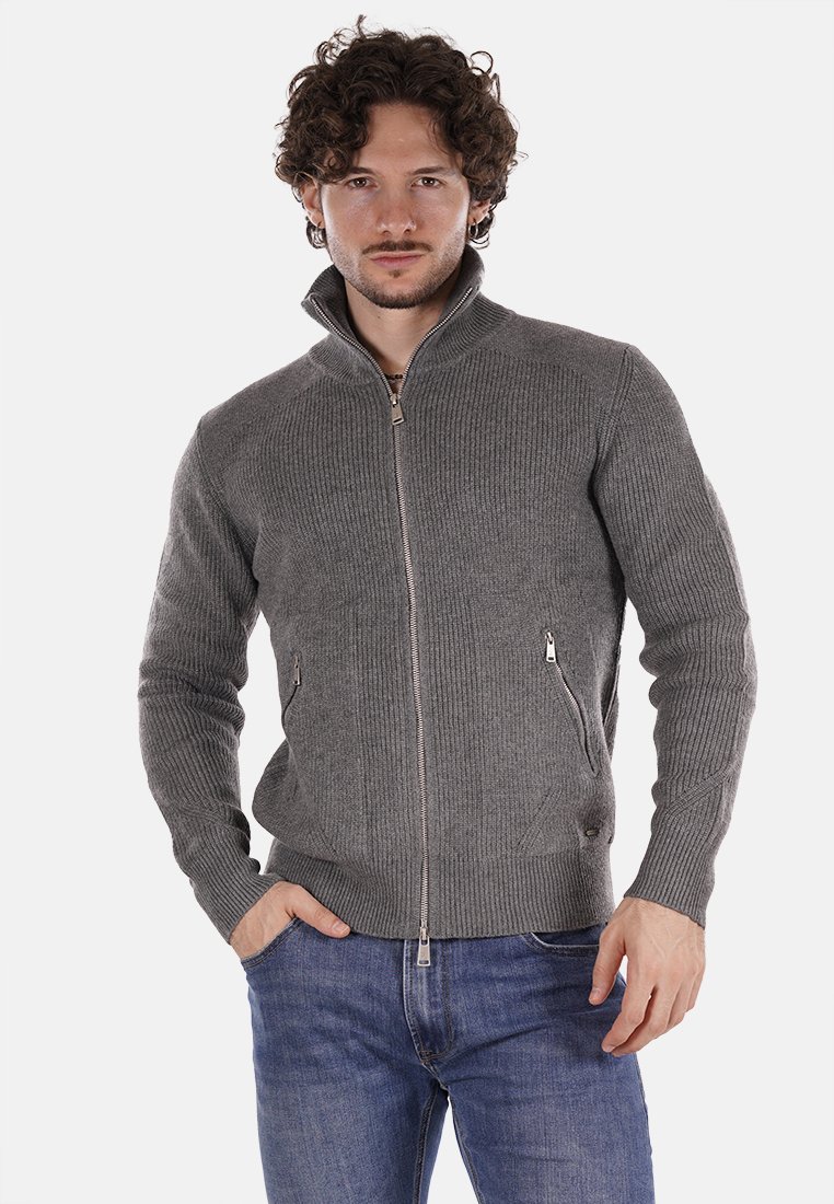 Maglione grigio lavorato a maglia con zip, texture a coste, colletto alto e tasche laterali, indossato sopra jeans in denim blu. Accenti argentati della zip.