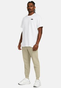 Camiseta blanca de algodón con un pequeño logo negro en el pecho, combinada con pantalones de chándal beige ajustados y zapatillas blancas.