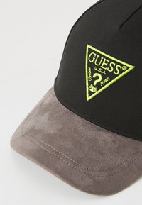 Casquette noire avec une visière en daim gris, comportant un logo triangulaire vert fluo portant l'inscription "GUESS" et un texte décoratif.