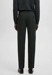 Osoba ubrana w czarne, dopasowane spodnie, czarne eleganckie buty oraz ciemnoszary sweter, stojąca tyłem na jednolitym tle.