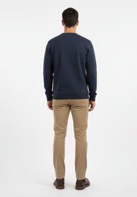 Sudadera azul marino con cuello redondo, puños y dobladillo acanalados; combinada con pantalones caqui y zapatos marrones. Vista trasera del conjunto.