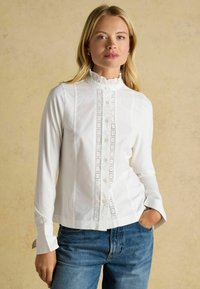 Weißes Langarmhemd mit Knopfleiste, das einen gerafften Kragen und filigrane Spitzendetails auf der Vorderseite aufweist. Kombiniert mit blauen Jeans.