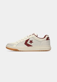 White leather sneakers with red accents, features a perforated toe cap, rubber sole, and a logo-decorated tongue. Smooth and textured details.
