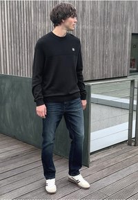 Svart tröja med en texturerad övre panel, blå jeans och vita sneakers. Står på en träplatta med en trävägg i bakgrunden.