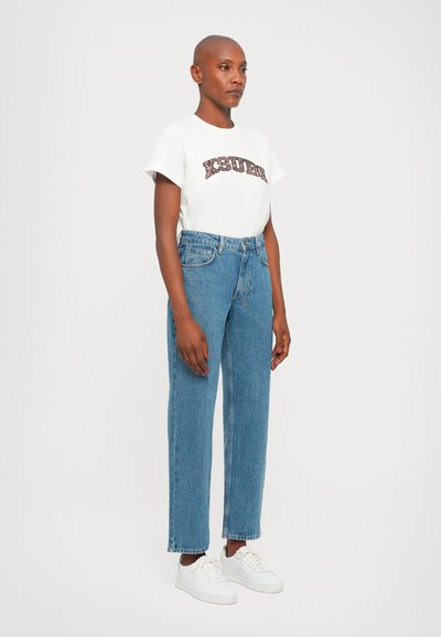 White t-shirt with a brown graphic print, paired with blue straight-leg jeans and white sneakers. Simple design with classic fit and fabric texture.