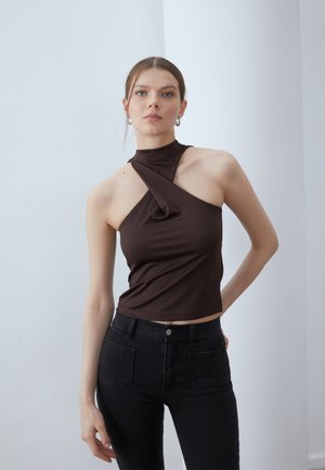 Un haut sans manches marron présente un col montant et un détail torsadé sur le devant. Associé à un jean noir taille haute. Texture lisse.