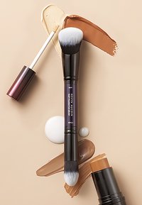 Doppelkopf-Make-up-Pinsel mit einer zugespitzten Kopf aus weißen und schwarzen Borsten. Umgebende flüssige Foundations in Beige und Braun auf einem cremigen Hintergrund.