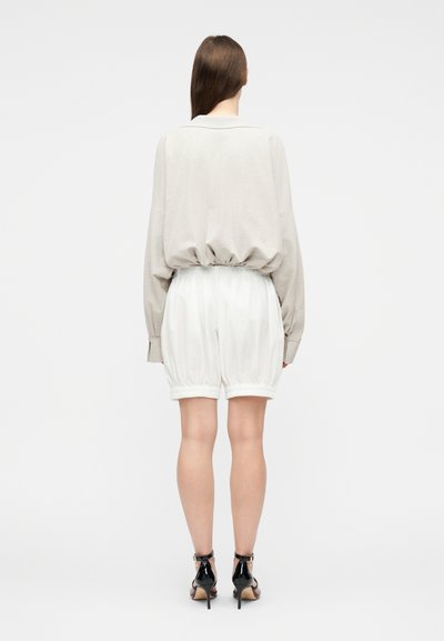 Γυναίκα που στέκεται με την πλάτη, φορώντας μπεζ μπλούζα με μακριά μανίκια και σούρα στη μέση, λευκά ελαστικά σορτς και μαύρα σανδάλια με ψηλό τακούνι.