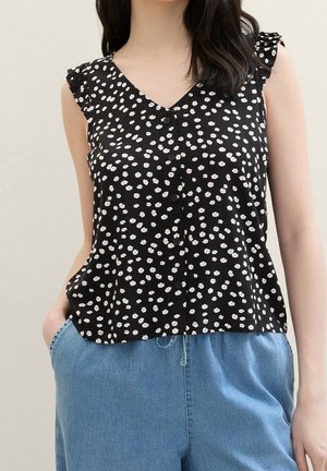 Mujer lleva una blusa negra sin mangas con estampado floral blanco y botones, combinada con pantalones vaqueros azules sueltos, posando con una mano en el bolsillo.