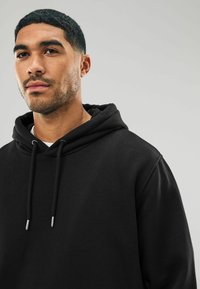 Schwarzer Hoodie mit lockerer Passform, der über eine Kordelzugkapuze, eine frontale Kängurutasche und eine weiche Textur verfügt. Silbereinfassungen an den Kordeln setzen Akzente.