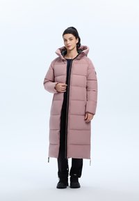 Roze pufferjas met capuchon, voorzien van een ritssluiting aan de voorkant en een verticaal gestikt patroon. Gestyled met zwarte broek en laarzen.