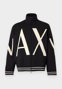 Veste noire zippée à col montant avec de grandes lettres crème "NAXY" sur le devant et les manches, avec poignets et bas rayés blancs.