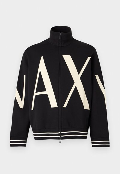 Veste noire zippée à col montant avec de grandes lettres crème "NAXY" sur le devant et les manches, avec poignets et bas rayés blancs.