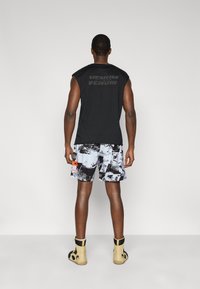 Μαύρο αμάνικο μπλουζάκι με το λογότυπο «VENUM» στην πλάτη, σε συνδυασμό με σορτς με σχέδια σε γκρι, μαύρο και πορτοκαλί αποχρώσεις, φορεμένα με μπεζ μπότες.