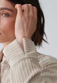 Tissu beige, côtelé et texturé avec des lignes verticales proéminentes. Le col présente un détail original avec un bord en forme de coquille.