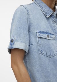 Chemise en denim bleu clair avec des poignets retroussés, deux poches poitrine et un col pointu. Le tissu a une texture légèrement délavée.
