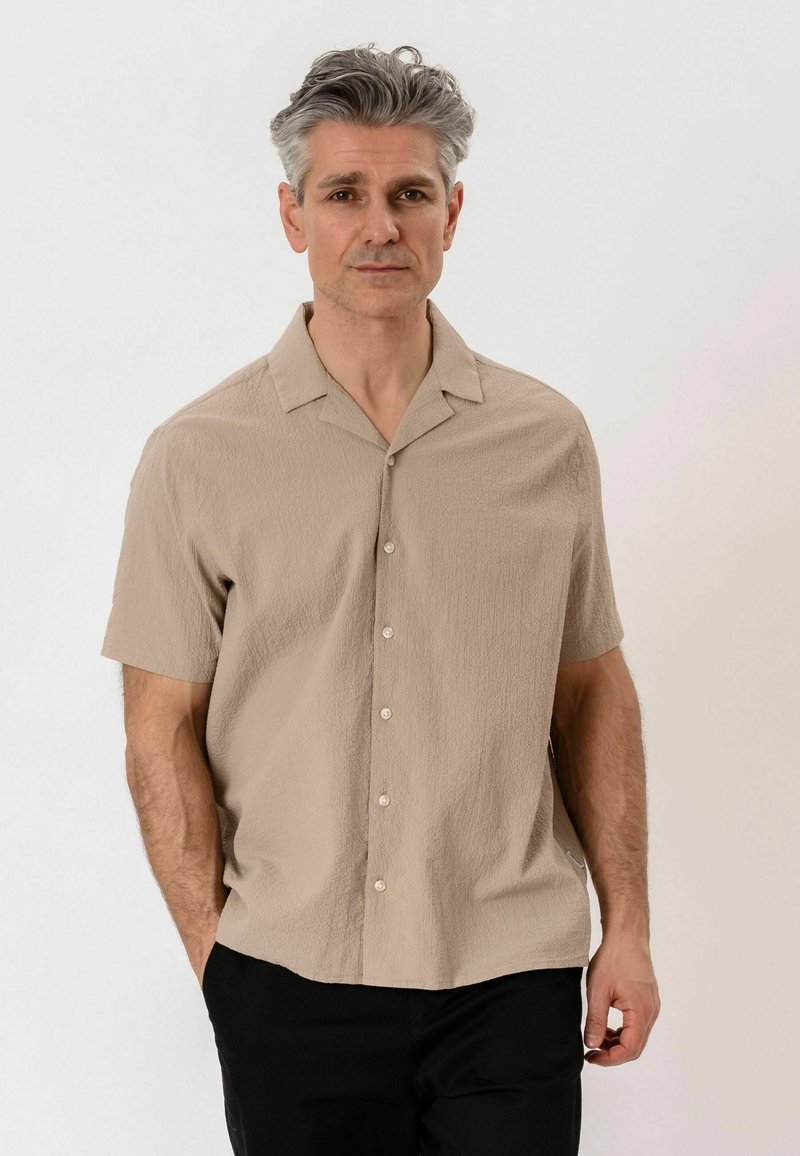 Homme aux cheveux gris portant une chemise beige à manches courtes boutonnée et un pantalon noir, debout les mains dans les poches sur un fond uni.