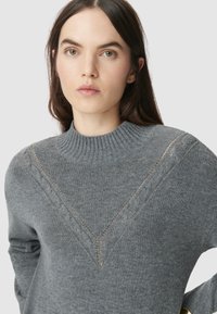 Szary sweter z dzianiny o prążkowanym wysokim dekolcie, z V-kształtnym wzorem i delikatnymi detalami w warkoczowym splocie wzdłuż dekoltu.