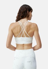 Reggiseno sportivo bianco con spalline incrociate e motivo marmorizzato. Dettaglio in tessuto testurizzato. Modella mostrata di spalle, abbinata a leggings coordinati.