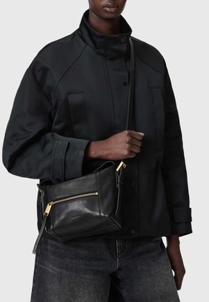 Personne portant une veste noire et un jean foncé, tenant un sac bandoulière en cuir noir avec une fermeture éclair dorée et des détails sur la bandoulière.