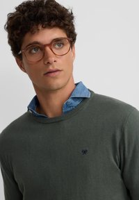 Suéter verde oliva con cuello redondo, superpuesto a una camisa de cuello clara azul. Gafas redondas con montura transparente y un pequeño detalle de logotipo en el suéter.