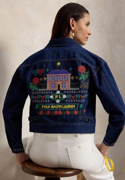 Femeie purtând o jachetă din denim albastru închis cu o casă brodată colorată, flori și textul „Polo Ralph Lauren” pe spate, stând pe un scaun înalt.