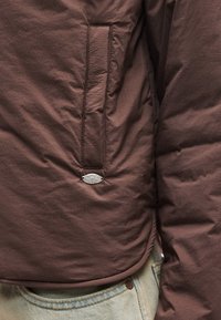 Καφέ μπουφάν puffer με λεία εξωτερική επιφάνεια, που διαθέτει πλαϊνή τσέπη και μεταλλική ετικέτα της μάρκας. Το τελείωμα καμπυλώνει πάνω από ανοιχτό μπλε τζιν.