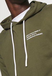 Olivenfarbene Kapuzenjacke aus Baumwollmischung, mit weißem Kordelzug, Kängurutasche und weißem Textbranding auf der Vorderseite.