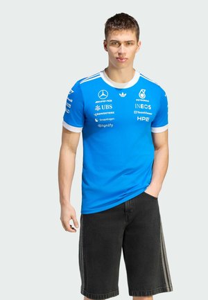 Giovane uomo che indossa una maglietta blu del team Mercedes-AMG Petronas con i loghi degli sponsor e pantaloncini neri, in piedi con una mano in tasca.