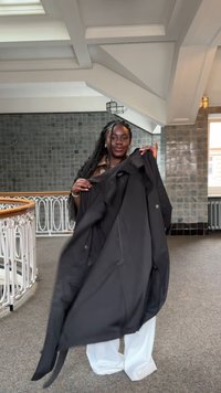 Czarny płaszcz o gładkiej teksturze, z długimi rękawami i zapięciem na guziki, trzymany otwarty na jasnym tle.