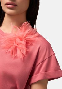 Roze t-shirt met gerolde mouwen heeft een grote, textuurvolle koraalstoffen bloem op de halslijn, wat zorgt voor dimensie en kleurcontrast.