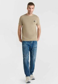 Camiseta de manga corta beige con un bolsillo en el pecho, combinada con jeans de denim azul con parches y múltiples bolsillos, y zapatillas grises.