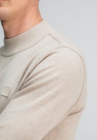 Beiger Pullover mit geripptem Kragen und strukturiertem Stoff, mit einem kleinen quadratischen Logo auf der linken Brust und eng anliegenden Ärmeln.