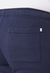 Tumed sinised dressipüksid sisaldavad nähtavat tagataskut, millel on silt. Kangas on pehme ja tekstureeritud, koos õmmeldud vöökohtadega.